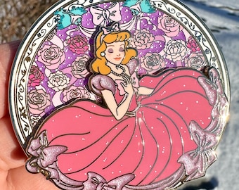 May include: A round, gold-toned pin featuring Cinderella in her iconic blue ball gown. The pin is surrounded by a floral design with pink roses and purple accents. The pin has a sparkly finish.