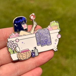 May include: A gold-tone enamel pin depicting a cartoon woman in a bathtub with a towel, bath products, and a bubble wand. The woman has blue hair and is wearing a pink bathrobe. The bathtub is white with pink and purple accents.