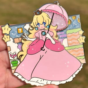 May include: A pink and gold enamel pin featuring a cartoon princess character holding a pink umbrella. The princess is wearing a pink dress with a white ruffle and a crown. The background is a pastel blue and pink landscape with stars and clouds.