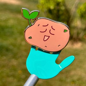 May include: A gold-tone enamel pin featuring a cartoon potato with a happy face and a green sprout growing from the top. The potato is sitting on a turquoise hand.