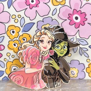 May include: Enamel pin depicting two cartoon characters, one in a pink dress and the other in a black witch hat. The background is a floral pattern with pink, yellow, and purple flowers.