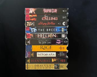 Może przedstawiać: Stos kaset VHS z tytułami horrorów, takimi jak "Vampire Clan", "The Calling" i "Sleepy Hollow". Kasety mają kolorowe okładki z tytułami i obrazami filmów. Stos znajduje się na czarnym tle.