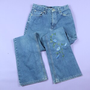 Puede incluir: Un par de pantalones vaqueros azules con un diseño de bordado floral en una pierna. Los vaqueros tienen un estilo de pierna recta y un lavado claro.