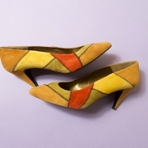 May include: A pair of high-heeled shoes with a patchwork design in shades of yellow, orange, and brown. The shoes are made of suede and have a pointed toe.