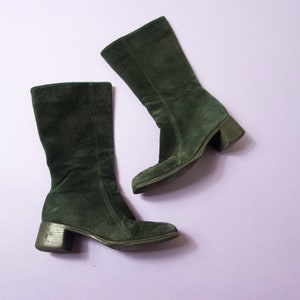 May include: Two dark green suede boots with a block heel are displayed against a light purple background. The boots are knee-high with a rounded toe and a zipper on the inner side. The heels are approximately 2 inches high.