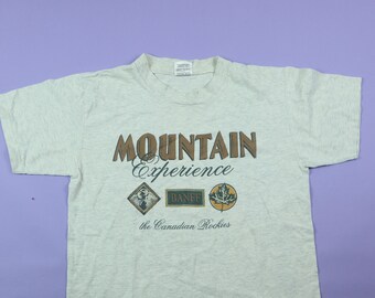 Könnte beinhalten: Ein hellgraues T-Shirt mit einer braunen Grafik, auf der "Mountain Experience" steht, darunter drei kleinere Grafiken. Die kleineren Grafiken zeigen eine Bergszene, das Wort "Banff" und ein Ahornblatt. Der Text unter den Grafiken lautet "The Canadian Rockies".