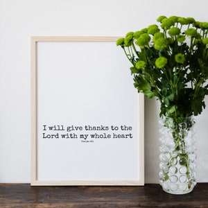 May include: A framed print with the text "I will give thanks to the Lord with my whole heart" in a simple black font. A clear glass vase with green flowers sits to the right of the frame. The frame is light wood.