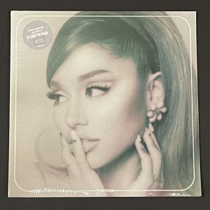 May include: A vinyl record cover featuring a close-up portrait of Ariana Grande. The cover is a light green color with a white border. The text "Positions" is printed in white at the bottom of the cover.