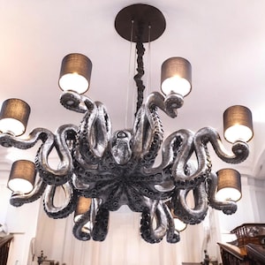 May include: A unique, octopus-shaped chandelier with eight lights. The fixture is silver-toned with black lampshades. The tentacles of the octopus curve and extend outward, creating a dramatic lighting fixture. The chandelier is suspended from the ceiling.