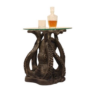 May include: A bronze-colored octopus-shaped side table with a round glass top. A decanter of amber liquid and a glass sit on the table. The table's base features detailed tentacles and a textured finish.