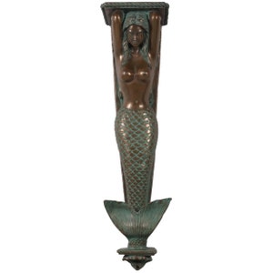 May include: Bronze-colored mermaid shelf bracket. The mermaid figure has long hair and holds a rectangular shelf support above her head. The mermaid's tail is detailed with scales and ends in a flared tail fin. The bracket has a verdigris finish.