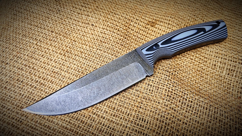 Op de afbeelding: Een mes met vast blad met een getextureerd, grijs lemmet en een handvat met een zwart-wit gestreept patroon. Het mes ligt op een getextureerde, bruine achtergrond.
