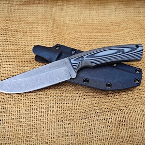 May include: A fixed-blade knife with a grey blade and a handle with a black and grey wood grain pattern. The knife is in a black sheath. The knife is on a brown burlap surface.