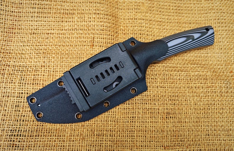 Op de afbeelding: Een zwart mes met een zwart-grijs gestreepte handgreep is vastgezet in een zwarte schede. De schede heeft een riemclip en is aan de handgreep bevestigd. Het mes en de schede liggen op een getextureerd, bruin oppervlak.