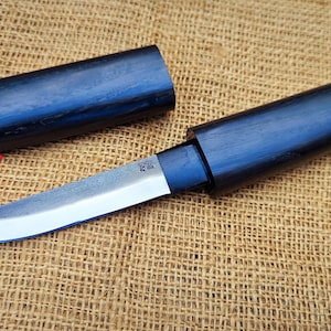 May include: A handcrafted knife with a polished steel blade and a dark blue wooden handle and sheath. The knife is partially removed from its sheath, showcasing the blade's edge. The wooden sheath is cylindrical.