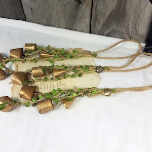 Op de afbeelding: Een set gouden bellen met groen gebladerte en touw, hangend aan een bruin touwkoord met twee bruine knopen.