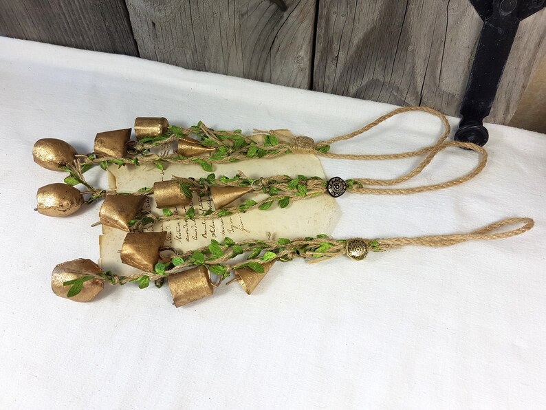 Op de afbeelding: Een set gouden bellen met groen gebladerte en touw, hangend aan een bruin touwkoord met twee bruine knopen.