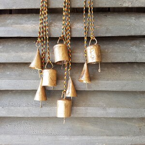 May include: A set of eight gold-coloured bells hanging from colourful cords against a wooden background. The bells are of various shapes and sizes, with some having a cone shape and others a more traditional bell shape.