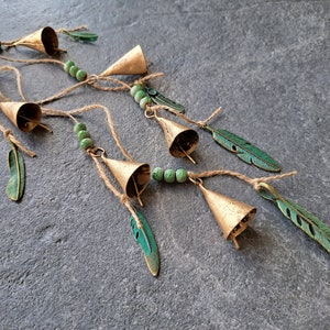 Op de afbeelding: Een reeks bellen met groene veren en kralen. De bellen zijn goudkleurig en de veren zijn donkergroen. De kralen zijn lichtgroen.