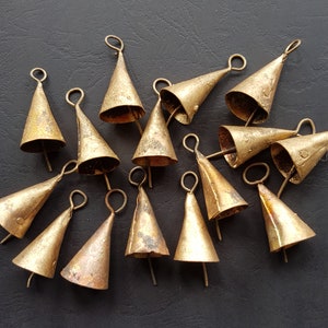 May include: A group of antique brass-coloured bells, each with a conical shape and a loop for hanging. The bells have small clappers and are arranged on a dark surface, highlighting their aged, textured appearance. The bells are made of brass.