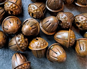 May include: A collection of small, bronze-colored bells. Each bell has a rounded top with a textured surface and vertical slits. The bells are scattered across a dark surface, creating a rustic and vintage aesthetic.