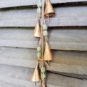 Op de afbeelding: Een reeks gouden bellen met groene kralen die aan een houten luik hangen. De bellen zijn in een cascade-achtige opstelling gerangschikt, met de grootste bel bovenaan en de kleinste bel onderaan.