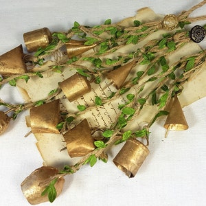 Op de afbeelding: Een guirlande van gouden belletjes met groene bladeren en touw. De belletjes zijn aan elkaar gebonden met touw en hebben een rustieke uitstraling.
