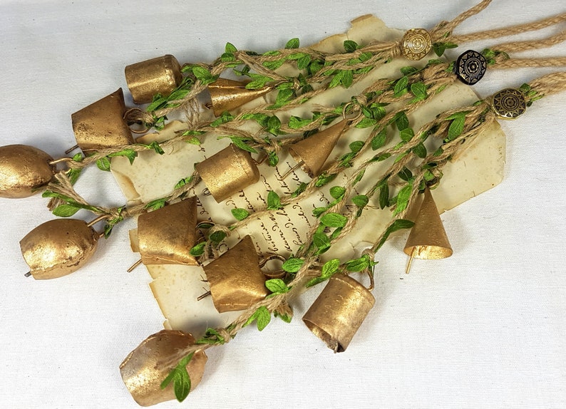 Op de afbeelding: Een guirlande van gouden belletjes met groene bladeren en touw. De belletjes zijn aan elkaar gebonden met touw en hebben een rustieke uitstraling.