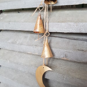 May include: A gold-colored wind chime with a crescent moon shape and multiple small bells hanging from it. The chime is hanging from a wooden surface.