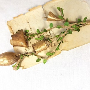 Peut inclure: Une chaîne de clochettes dorées avec des feuilles vertes attachées à un morceau de ficelle. Les clochettes sont suspendues à un morceau de vieux papier avec de l'écriture manuscrite.