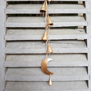 May include: A gold metal crescent moon wind chime with a series of small, gold bells hanging from it. The chime is hanging from a wooden shutter.