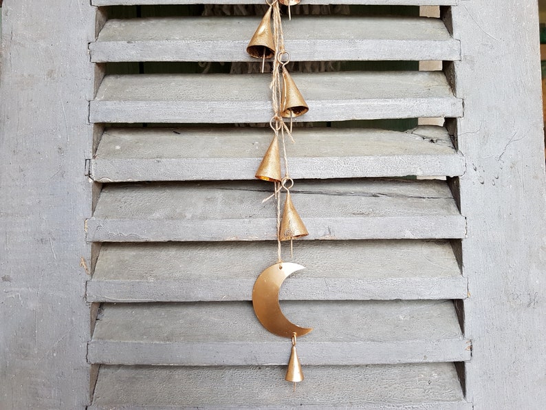 May include: A gold metal crescent moon wind chime with a series of small, gold bells hanging from it. The chime is hanging from a wooden shutter.