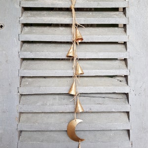 May include: A gold metal crescent moon with a string of small gold bells hanging from it. The bells are attached to a twine string and the moon is hanging from a wooden shutter.