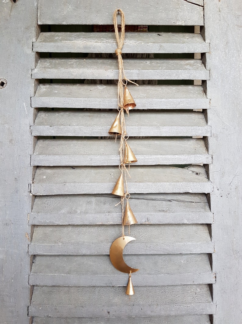 May include: A gold metal crescent moon with a string of small gold bells hanging from it. The bells are attached to a twine string and the moon is hanging from a wooden shutter.