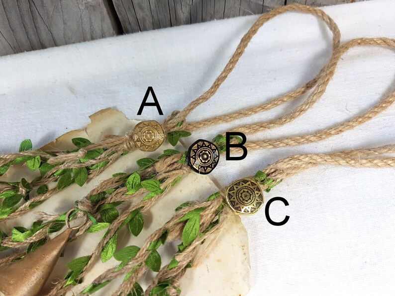 Op de afbeelding: Drie bruine jute koorden met decoratieve metalen bedels. De bedels zijn goudkleurig met een zwart centrum en een geometrisch ontwerp. De koorden zijn gemarkeerd met A, B en C.