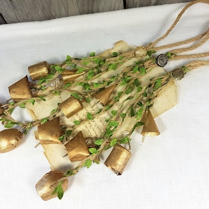 Op de afbeelding: Een guirlande van gouden belletjes met groene bladeren en touw, vastgemaakt met twee decoratieve knopen. De guirlande hangt aan een wit oppervlak.