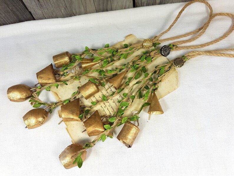 Op de afbeelding: Een guirlande van gouden belletjes met groene bladeren en touw, vastgemaakt met twee decoratieve knopen. De guirlande hangt aan een wit oppervlak.
