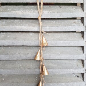 May include: A gold-colored metal wind chime with five bells hanging from a length of twine. The bells are cone-shaped and have a rustic, aged look.