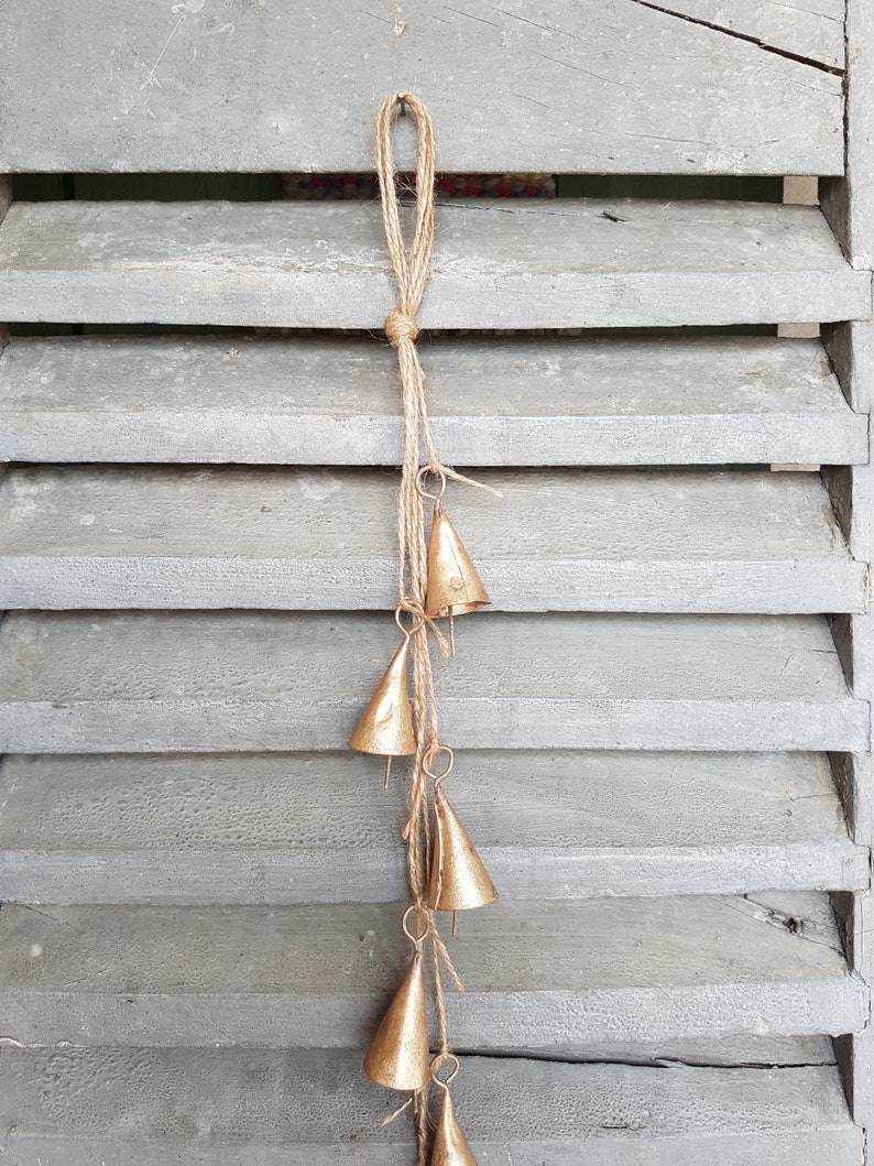 May include: A gold-colored metal wind chime with five bells hanging from a length of twine. The bells are cone-shaped and have a rustic, aged look.