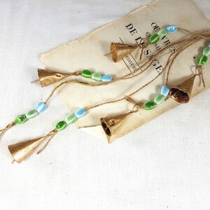 Op de afbeelding: Een set van vijf gouden bellen met groene en blauwe glazen kralen die aan een touwtje zijn geregen. De bellen hangen aan een boek met de zichtbare tekst "DE L'SAGE".