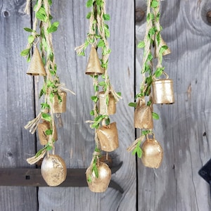 Op de afbeelding: Drie strengen gouden bellen die aan een houten oppervlak hangen. Elke streng is versierd met groene bladeren en bruin touw. De bellen zijn van verschillende groottes en vormen.