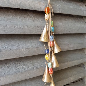 Op de afbeelding: Een reeks bellen met kleurrijke kralen die aan een houten oppervlak hangen. De bellen zijn goud en de kralen zijn rood, oranje, geel, blauw en groen.
