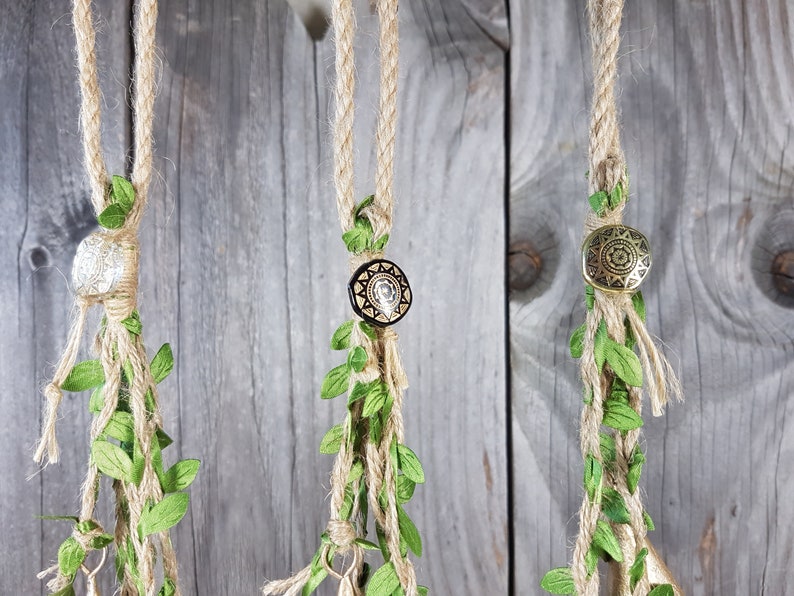 Op de afbeelding: Drie macram&eacute; plantenhangers met verschillende gekleurde kralen en groene bladeren die aan een houten muur hangen.