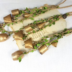 Op de afbeelding: Een reeks gouden belletjes met groene bladeren en touw, hangend aan een rustiek bruin touw. De belletjes zijn aan elkaar gebonden met touw en hebben een rustieke, vintage uitstraling.