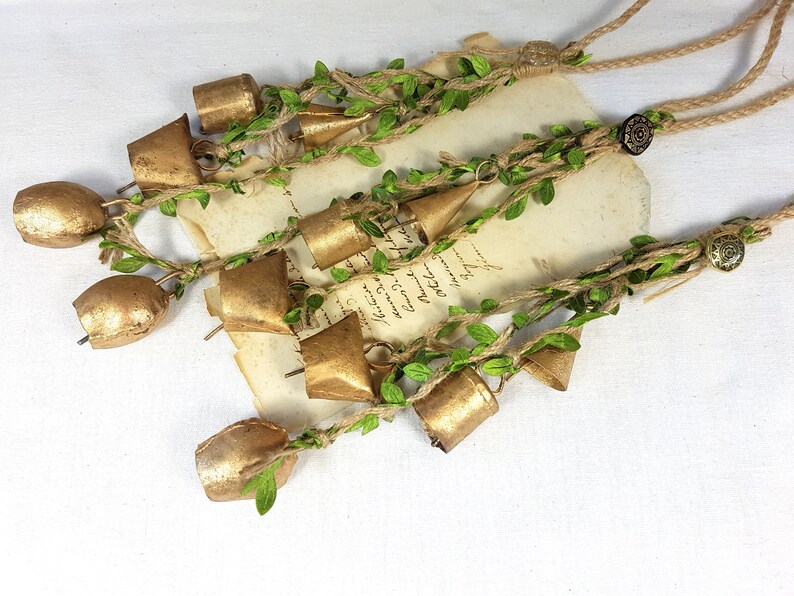 Op de afbeelding: Een reeks gouden belletjes met groene bladeren en touw, hangend aan een rustiek bruin touw. De belletjes zijn aan elkaar gebonden met touw en hebben een rustieke, vintage uitstraling.