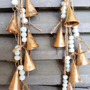 May include: A gold-coloured metal bell garland with white and light blue beads hanging from a wooden surface. The bells are arranged in a cascading pattern, creating a whimsical and festive look.