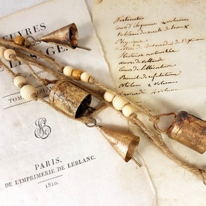 May include: A string of aged metal bells with a rustic finish, interspersed with wooden beads, hangs against a backdrop of old paper. The bells are cone-shaped and cylindrical. The paper has handwritten text and the text "PARIS, DE L'IMPRIMERIE DE LEBLANC. 1810."