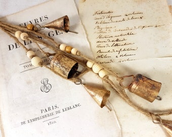 May include: Rustic metal bells with a distressed finish are strung on twine, interspersed with wooden beads. The bells are cone-shaped and cylindrical. The background includes aged paper with handwritten text and the text "PARIS, DE L'IMPRIMERIE DE LEBLANC. 1810."
