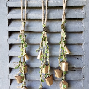 Op de afbeelding: Drie gouden bel-slinger met groene bladeren die aan een houten luik hangen. Elke slinger heeft een klein, rond, gouden sieraad aan de bovenkant.