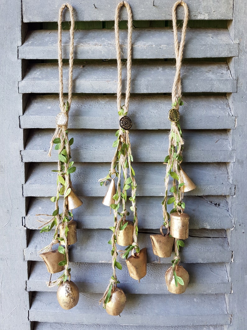 Op de afbeelding: Drie gouden bel-slinger met groene bladeren die aan een houten luik hangen. Elke slinger heeft een klein, rond, gouden sieraad aan de bovenkant.